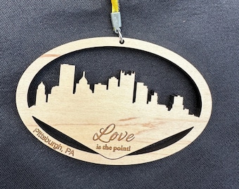 May include: Wooden ornament shaped like an oval with a cutout of the Pittsburgh skyline. The ornament is painted brown and has the text "Love is the point!" and "Pittsburgh, PA" on it.