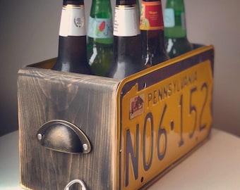 May include: A wooden six-pack beer carrier with a Pennsylvania license plate design. The license plate reads "PENNSYLVANIA NO.6.152". The carrier is filled with six bottles of beer.