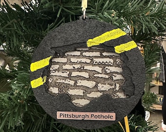 May include: A black and yellow Christmas ornament shaped like a pothole with a brick texture. The ornament has a brown tag that reads "Pittsburgh Pothole".