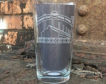 May include: A clear glass pint glass with a white etched design of the Homestead Steel Works. The design includes the words "Homestead" and "USS Steel Works".