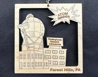 May include: A wooden ornament featuring a black and white line drawing of a building with a large dome. The building is labeled "Westinghouse Research Laboratories" and "Forest Hills, PA". A speech bubble above the building reads "ATOM SMASHER!"