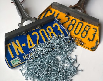 May include: Two vintage license plates, one blue and yellow with the text "TN-42083" and the other yellow with the text "CH 28083", are attached to two metal engine connecting rods. A pile of silver screws is spilling out from the bottom of the plates.