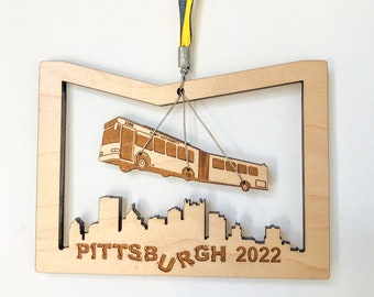 May include: Wooden ornament featuring a bus suspended above a cityscape silhouette. The ornament is light brown with the text "PITTSBURGH 2022" at the bottom. A yellow and blue ribbon is attached for hanging.