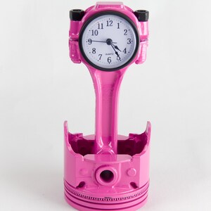 Op de afbeelding: Een roze motorzuigerklok met een witte wijzerplaat en zwarte wijzers. De klok is gemonteerd op een roze zuiger en staat op een roze zuigerbasis.