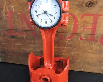 May include: A red metal clock shaped like a car engine piston. The clock face is white with black numbers and hands. The clock is set against a wooden background.
