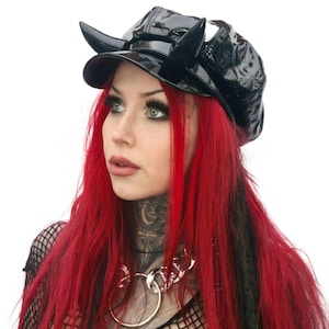 May include: A black patent leather newsboy cap with two curved horns. The hat has a shiny finish and a short brim. The person in the image has bright red hair and is wearing a silver chain necklace.