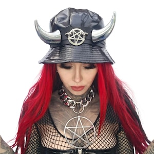May include: Black bucket hat with silver horns and a pentagram emblem. The hat is made of a shiny material. The hat is worn with a silver chain necklace with a large pentagram pendant.