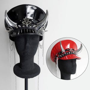 Puede incluir: Dos sombreros negros y rojos con cuernos plateados y cadenas. Los sombreros están en maniquíes con cabezas negras. Los sombreros tienen un diseño de pentagrama en la parte delantera.