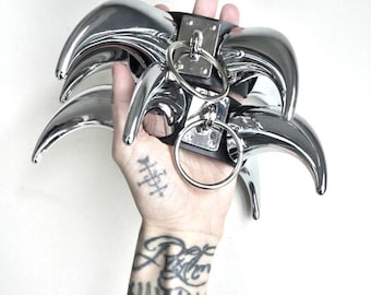 Puede incluir: Un conjunto de garras de metal plateado pulido con una correa de cuero negro y dos anillos plateados. Las garras se sostienen en una mano sobre un fondo blanco. Las garras tienen un diseño curvo y puntiagudo.
