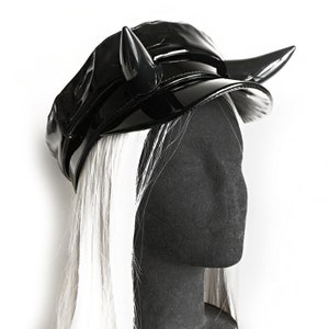 May include: A black, glossy, vinyl-like hat with two curved, horn-shaped accents. The hat has a visor and is displayed on a black mannequin head with long, white hair, against a white background.
