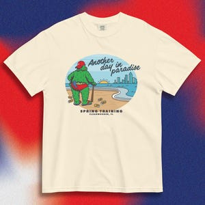 May include: Cream-colored t-shirt featuring a cartoon illustration. The graphic depicts a green, furry creature in red swim trunks and a baseball cap, standing on a beach with the text "Another day in paradise" and "Spring Training Clearwater, FL".