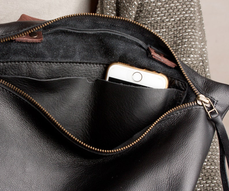 Op de afbeelding: Een zwarte leren ritszak met een bruine leren klep en een gouden rits. Een smartphone is zichtbaar in de tas.