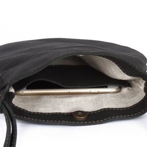 以下が含まれることがあります： フラップ留めと茶色のボタンが付いた黒い革製のショルダーバッグ。バッグが開いており、スマートフォンが中に入っているのが見えます。
