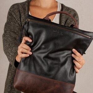 Op de afbeelding: Een zwarte leren tote bag met een bruine leren bies. De tas heeft een ritssluiting en een klein leren label met het woord "indigo" erop.