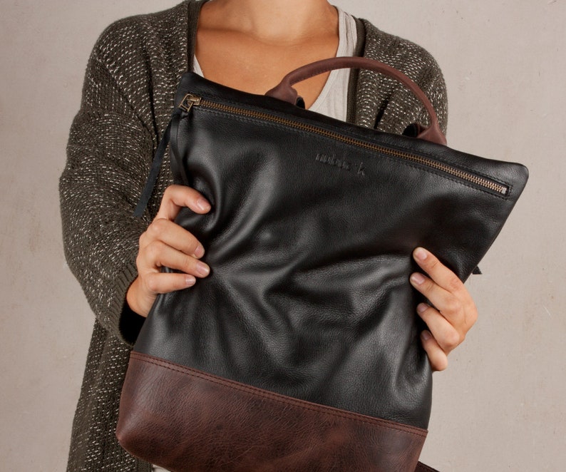 Op de afbeelding: Een zwarte leren tote bag met een bruine leren bies. De tas heeft een ritssluiting en een klein leren label met het woord "indigo" erop.