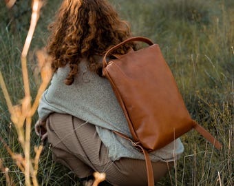 Puede incluir: Una persona con un suéter marrón y pantalones marrones está sentada en un campo de hierba alta. Lleva una mochila de cuero marrón con una correa.