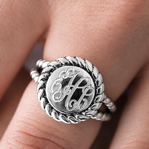 May include: A silver-toned ring featuring a circular face with an engraved monogram of the letters H, C, and B in a script font. The circular face is surrounded by a twisted rope-style border, and the band consists of two thin, twisted metal strands.