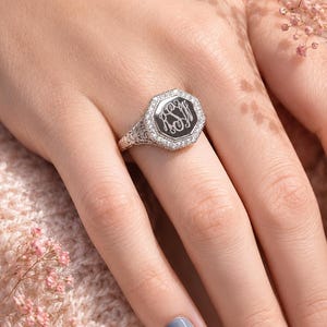 May include: A silver-toned octagonal signet ring featuring a central black inlay engraved with a decorative monogram. The ring face is bordered by a halo of small clear stones, and the band displays intricate filigree metalwork detailing.