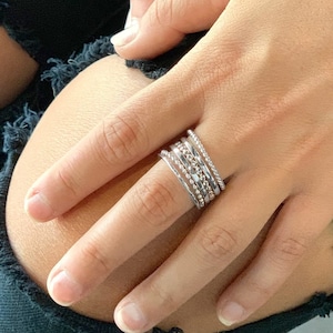 May include: A hand wearing a stack of five silver rings. The rings are different widths and have various textures, including a beaded ring and a twisted ring.