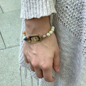 May include: A bracelet with beads in shades of beige, brown, and blue. It features a gold-coloured rectangular plaque with the initials "N M S" in a decorative script. The bracelet is worn on a wrist, with a knitted sleeve visible in the background.
