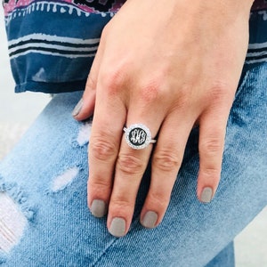 May include: A silver ring with a round black stone featuring a monogram. The ring is worn on the middle finger of a hand wearing light gray nail polish.