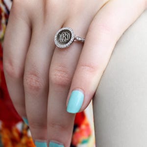 Könnte beinhalten: Ein silberfarbener Ring mit einer runden Platte, die in der Mitte mit dem Monogramm MBD graviert ist. Die runde Platte ist von einem Kranz aus kleinen, klaren, funkelnden Steinen umgeben, und die Ringschiene ist mit weiteren kleinen Steinen besetzt.