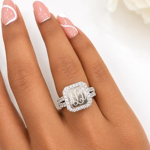May include: A silver-toned ring featuring a square-shaped face with a polished center engraved with the letters BLD in a cursive script. The square face is surrounded by a halo of small clear stones, and the band is also set with clear stones.