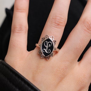 May include: A silver ring with a black oval stone featuring a white engraved letter L in a decorative filigree setting.