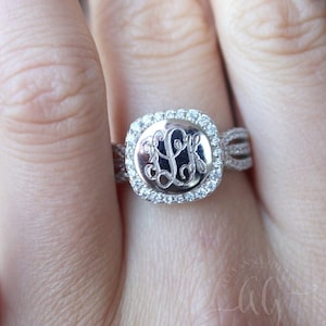 May include: A silver ring with a square-shaped center stone. The center stone is surrounded by smaller stones and features a monogrammed design. The ring band is a braided design.
