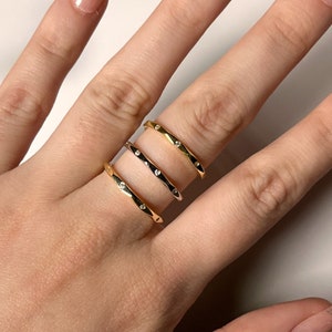 Puede incluir: Un conjunto de cuatro anillos dorados, cada uno con una banda delgada y pequeñas gemas transparentes. Los anillos están apilados en un dedo, mostrando su diseño. El fondo es de un color claro y neutro.