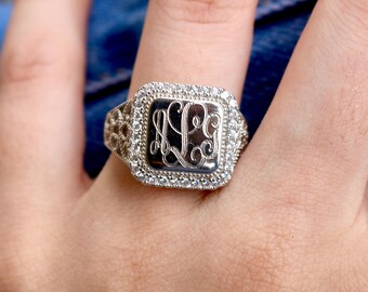 May include: A silver-toned ring featuring a square-shaped face with an engraved monogram in the center. The face is surrounded by a halo of small, clear stones, and the band displays an intricate, openwork filigree pattern on the sides.