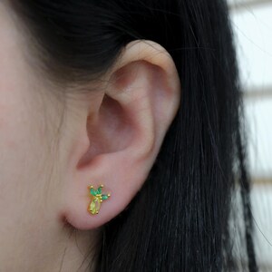 Puede incluir: Un pendiente de botón dorado con diseño de piña. El pendiente presenta una piedra amarilla de forma ovalada en la base, con piedras verdes que forman la parte superior. El pendiente está colocado sobre el fondo de una oreja y cabello oscuro.