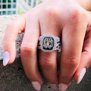 Puede incluir: Un anillo de plata con una piedra central de forma cuadrada que presenta un monograma de las letras "EHO". El anillo está engastado con pequeñas piedras transparentes alrededor de la piedra central y la banda.