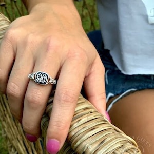 May include: A silver ring with a black square stone featuring a monogram. The ring is worn on the index finger of a person's hand.