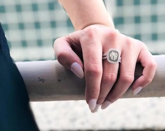 May include: A silver ring with a square-shaped center stone featuring a monogram. The ring is worn on the ring finger of a hand.