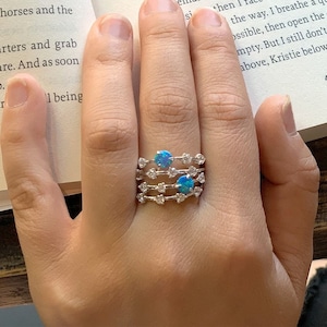 May include: A hand wearing a set of three silver rings with blue opal gemstones. The rings are stacked on top of each other and feature a delicate design with small white gemstones.