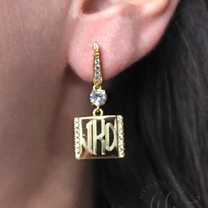 May include: Gold dangle earrings with a square charm featuring a monogram and clear rhinestones around the edge. The charm is attached to a small clear rhinestone.