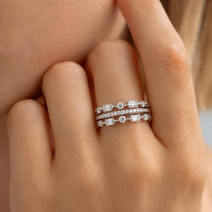 May include: A stack of three silver-toned rings worn on a finger. The top and bottom rings feature alternating baguette-cut and round-cut clear stones in a delicate metal setting. The middle ring is a thin band encrusted with small round stones.