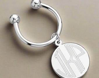 May include: A silver-colored key ring with a circular pendant. The pendant features an engraved monogram design. The key ring has a curved, horseshoe-like shape with a ball at each end. The pendant hangs from the ring.