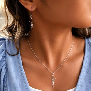 May include: A silver necklace and earrings set featuring a cross pendant and earrings. The word "Jesus" is written vertically on each piece. The jewelry is displayed against a blue background.