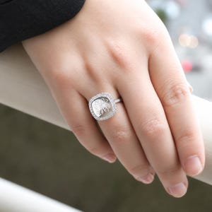 Könnte beinhalten: Ein silberfarbener Ring mit quadratischer Front und abgerundeten Ecken. In der Mitte des Rings befindet sich ein eingraviertes Monogramm, das von einem Kranz aus kleinen, klaren, funkelnden Steinen am äußeren Rand des Rahmens umgeben ist.