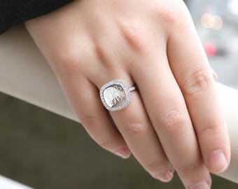 May include: A silver-toned ring featuring a square-shaped face with rounded corners. The center of the ring displays an engraved monogram, surrounded by a halo of small, clear, sparkling stones set along the outer edge of the square frame.