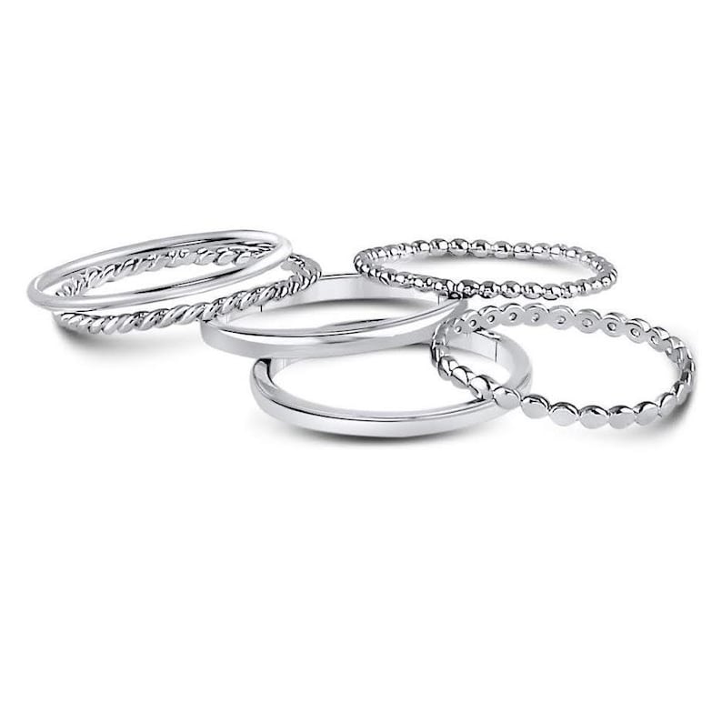 Puede incluir: Un conjunto de cinco anillos de tono plateado con diversas texturas, incluyendo bandas lisas, un dise&ntilde;o de cuerda trenzada y una banda con cuentas. Los anillos est&aacute;n dispuestos en grupo, mostrando su acabado met&aacute;lico y patrones.