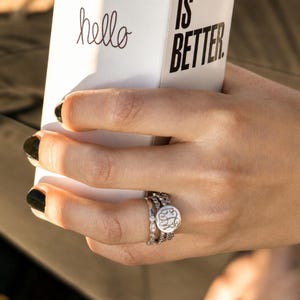 May include: A set of three silver-toned rings worn on a finger, including one band with a round monogrammed disc and two bands featuring small clear stones. The rings are held against a white box with the black text hello IS BETTER.