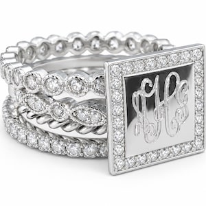 May include: A stack of four silver-toned rings featuring a square plaque with an engraved monogram. The set includes bands with round clear stones, a twisted metal texture, and a marquise-shaped stone detail surrounding the central square piece.