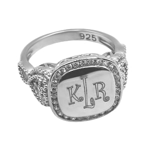 May include: A silver ring with a square-shaped center stone set with small clear stones. The center stone is engraved with the initials "KLR". The ring is marked "925" on the inside band.