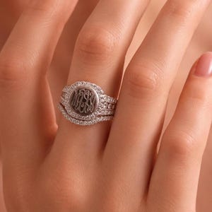Könnte beinhalten: Ein silberfarbenes Ring-Set mit einer zentralen runden Scheibe, die mit einem dreibuchstabigen Monogramm graviert ist. Die Scheibe ist von einem Kranz aus kleinen klaren Steinen umgeben, ergänzt durch weitere Steine am Ringband.