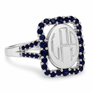 May include: A silver-toned ring featuring a central oval plate engraved with a three-letter monogram. The oval is framed by a rectangular border of small, round, deep blue stones. The split shank is also accented with rows of matching blue stones.
