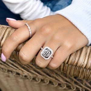 Könnte beinhalten: Ein silberfarbener Ring mit einer rechteckigen Platte mit dunkler Mitte und den Monogrammbuchstaben MYA, umgeben von einem Kranz aus kleinen klaren Steinen. Ein zweiter dünner, zierlicher silberfarbener Ring mit kleinen klaren Steinen am Zeigefinger.