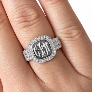 May include: A silver-toned ring featuring a square-shaped center with rounded corners, inlaid with a black surface displaying the engraved monogram letters GLH. The center is surrounded by a halo of small clear stones, with additional stones set along the band.